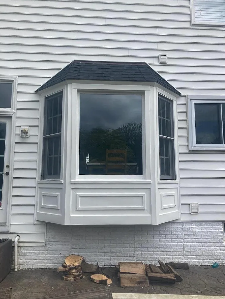 White bay window with a shingled roof cap installed on a light grey sided home